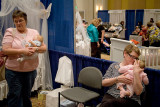 Baby Love : Mothers sharing their artificial infants at a convention.  In the background are displays of babies presented for adoption.