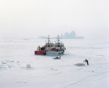Allanngorpoq 2 : An employee unloads boxes onto a sled in front of two boats trapped in the ice - Over the last few decades, Greenland’s society has undergone profound evolution. Thus, as the environment shifts, its people begin to embrace Western lifestyles and modes of consumption in parallel. Supermarkets, churches and cell phones are slowly making their way into Inuit culture. For the teenagers in the major towns the memories of seal hunting trips are long gone. And when they still occur in the northernmost dwellings the traditional outfits made from animal hides mix with modern fabrics, boats are used in combination with sledges.  These radical and rapid changes raise questions about society and identity, and divide public opinion in Greenland. Its people are torn between a desire to catch up with the modern world, and a feeling that they are an ice population which, like the ice itself, is slowly melting away.  “Allanngorpoq” in Greenlandic can be translated as “being...